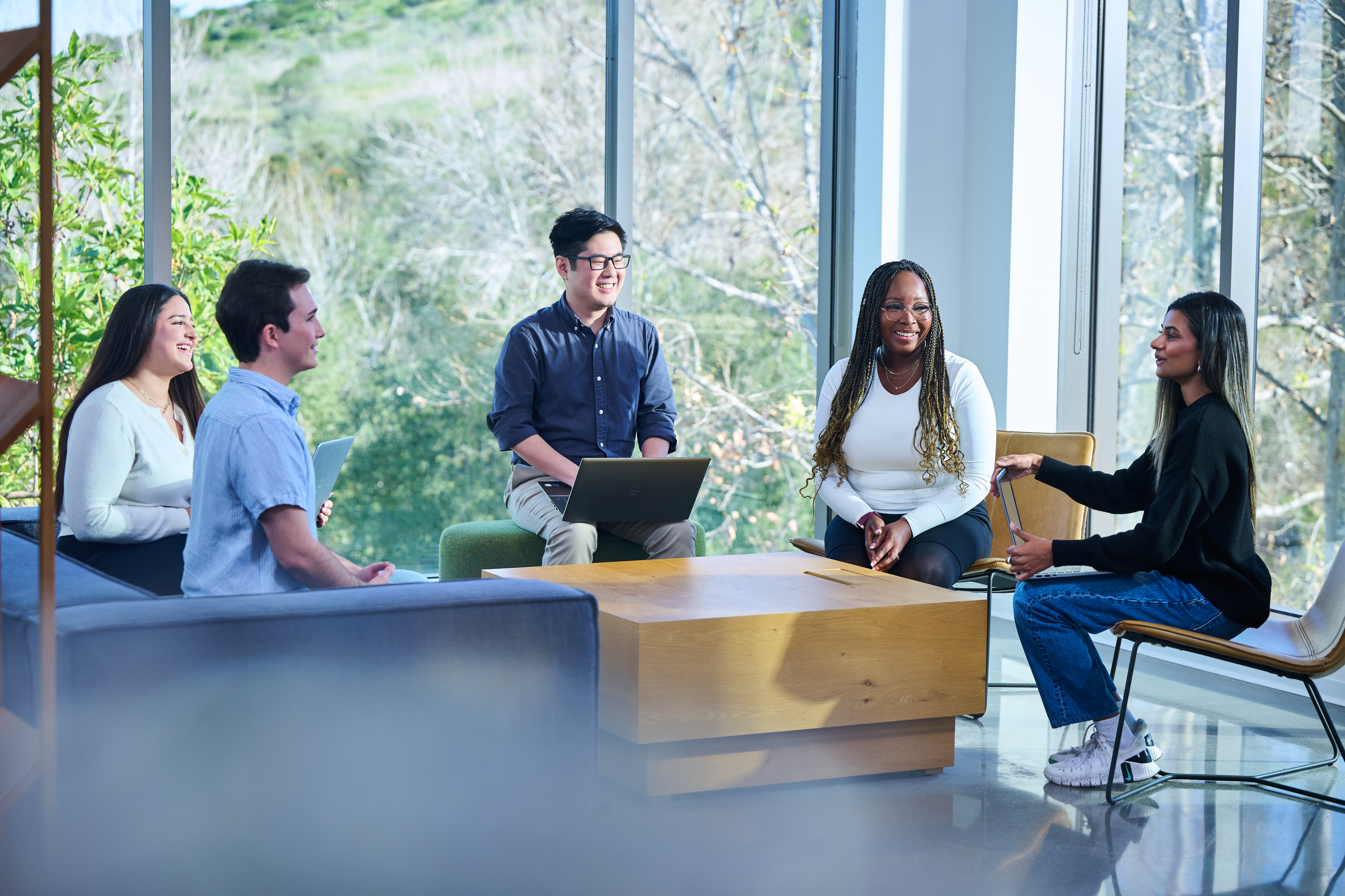 Five people engage in conversation around a wooden table in a bright, modern office space.