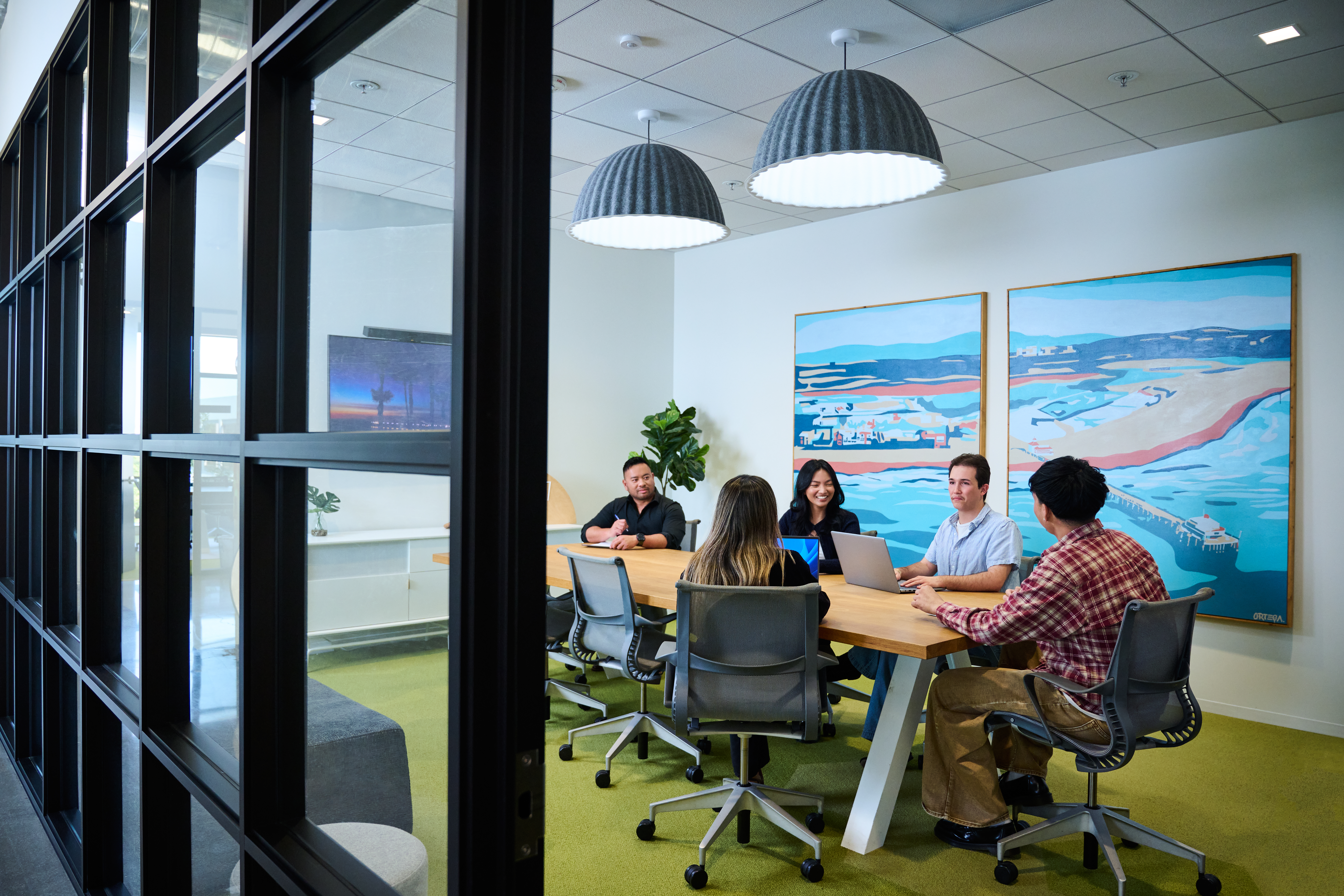 A group of five people collaborates around a conference table in a modern office. Colorful artwork decorates the walls.