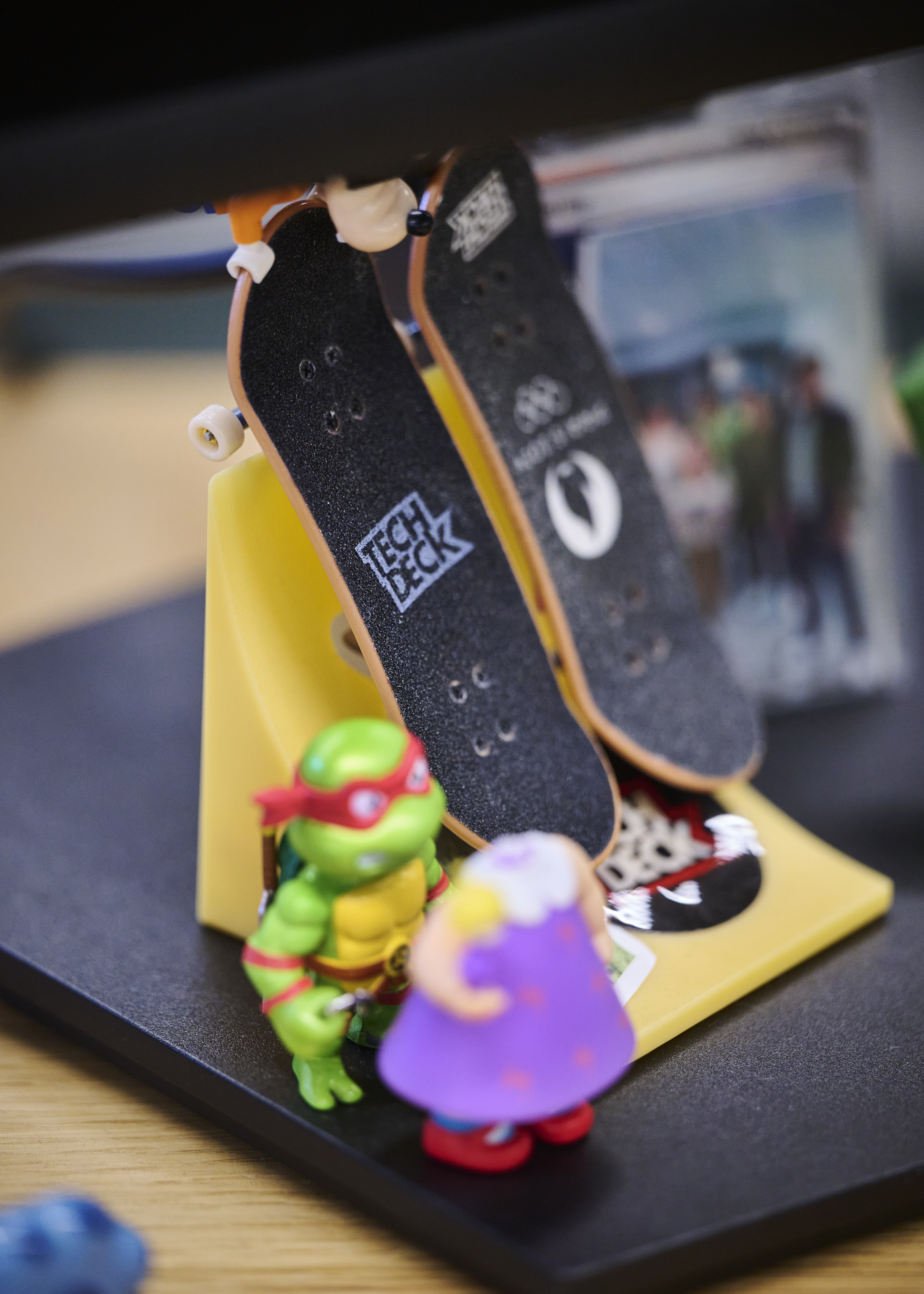 Two miniature skateboards on a stand, with a green turtle figure and a small character in a purple dress nearby.