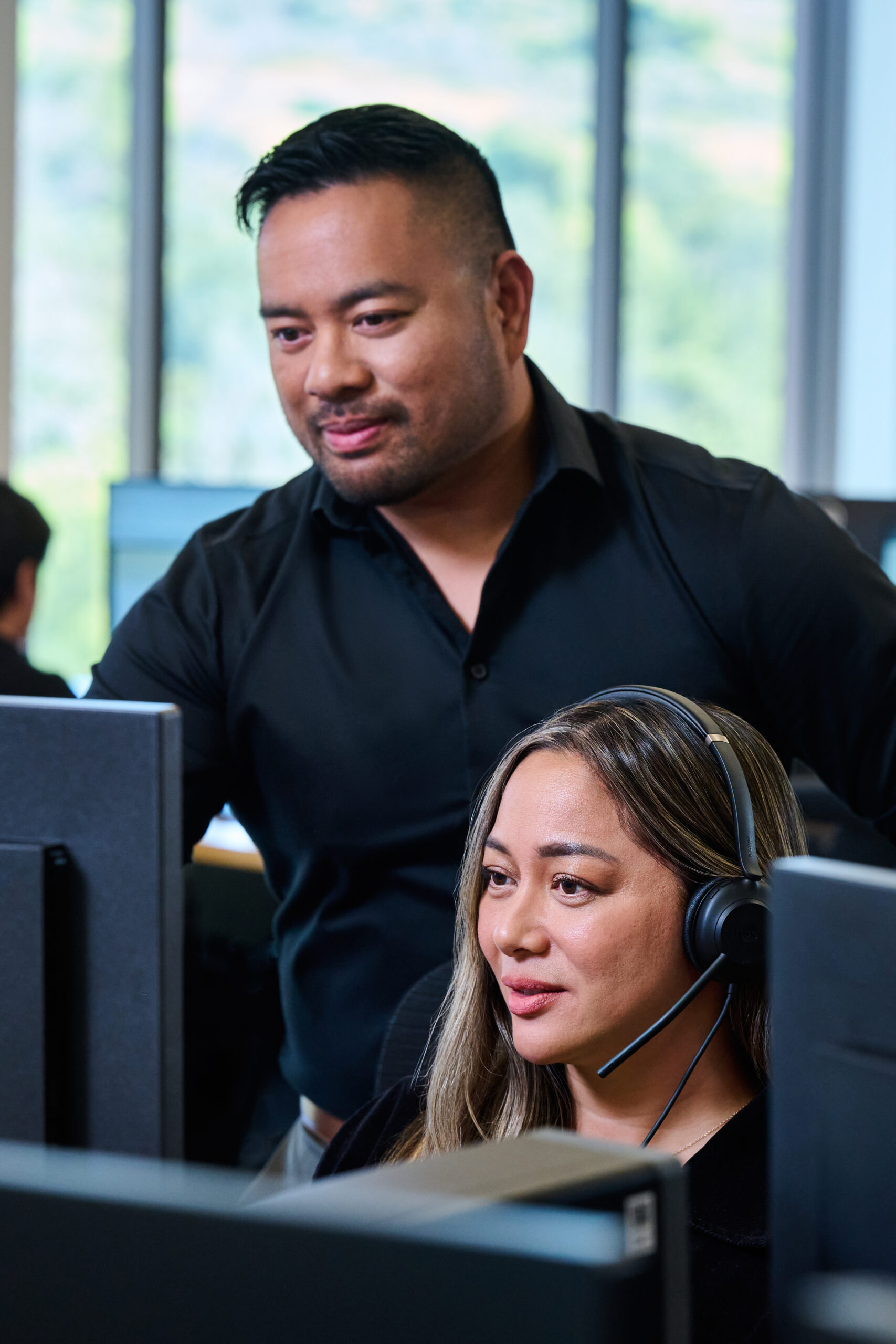 A man stands behind a woman at a computer, providing assistance while she wears a headset.